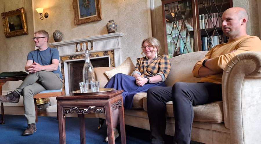 Man sitting on chair interviewing a man and a woman sitting on a sofa nearby.
