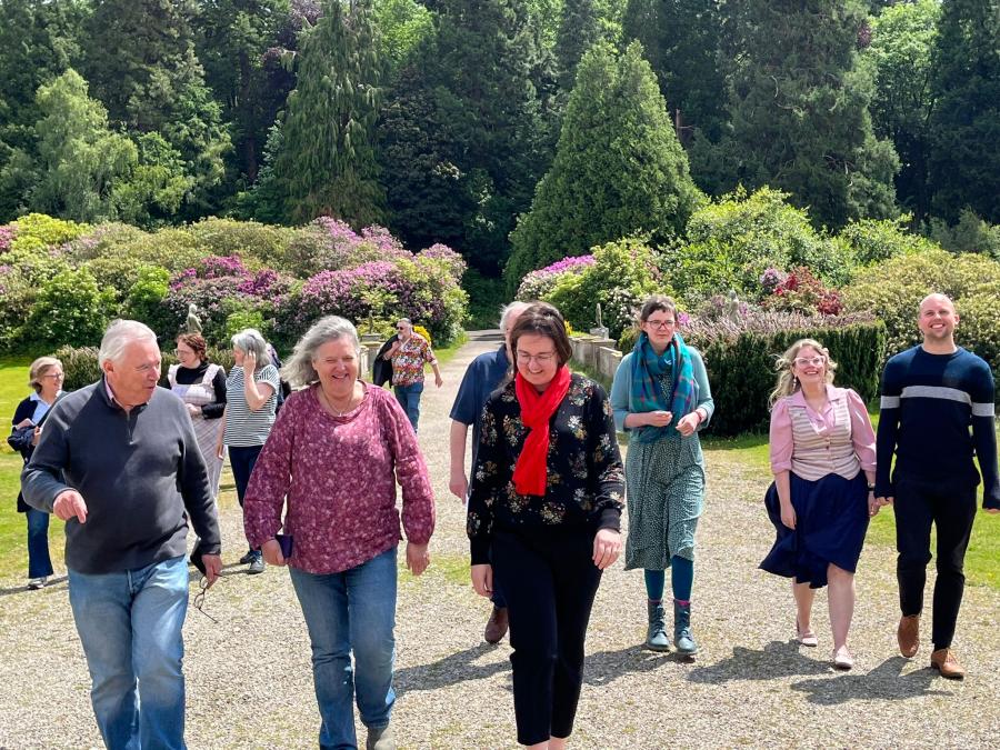 A group of people walking, all in conversation and smiling. Background of trees and flowering shrubs.