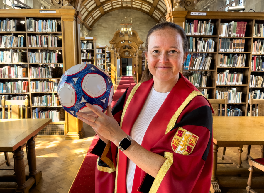 Llun o Cheryl Foster mewn gŵn graddio coch ac aur yn dal pêl droed