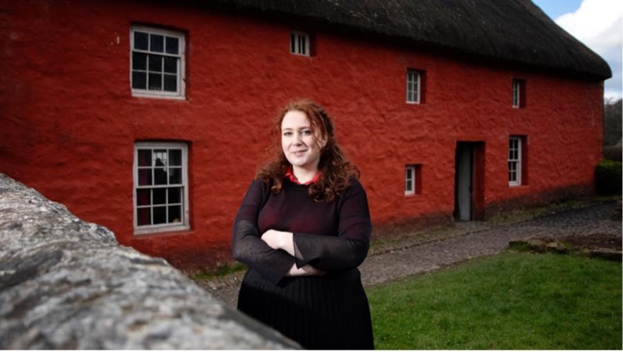 Bethan wears black and stands in front of a red farmhouse