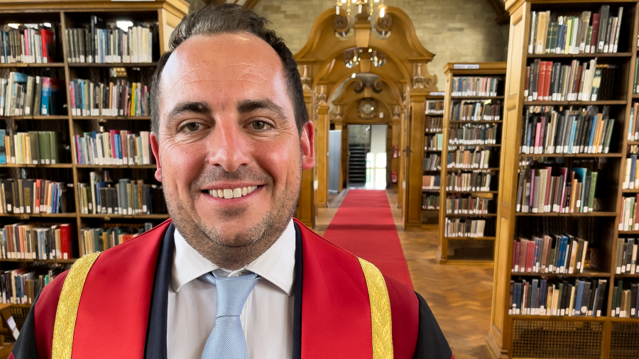 Llun o Toby Dixon mewn gŵn graddio coch ac aur