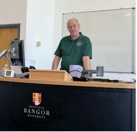 Vic wears a green ISWE polo shirt and stands behind a lectern 