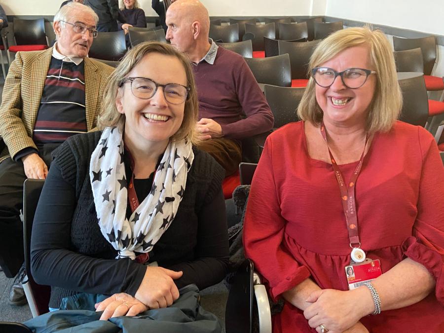 Two women smiling in the foreground, while two men are talking in the background.