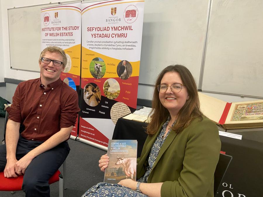 Dr Shaun Evans sits on the left, in front of the ISWE banners. Dr Lowri Ann Rees sits on the right, holding a copy of Shaun's book.