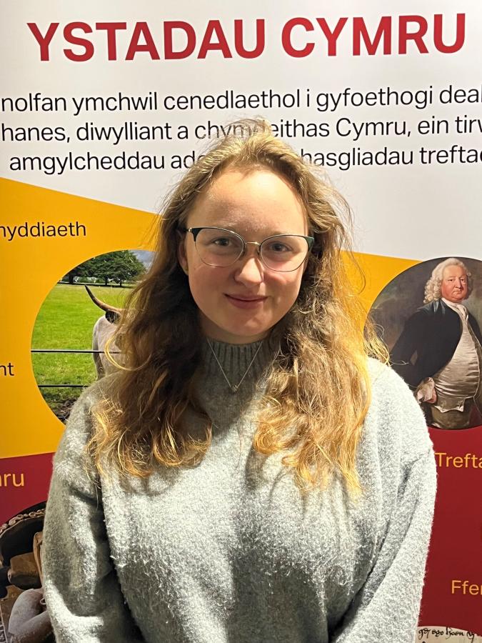 Isabel, who is wearing a grey jumper, is smiling and standing in front of an ISWE banner
