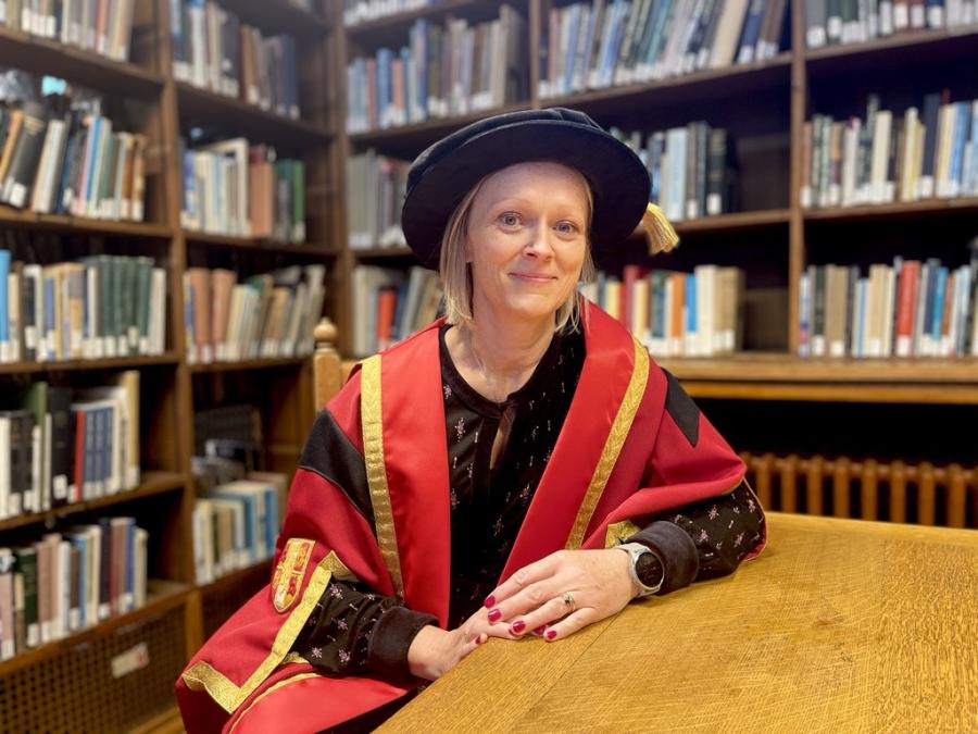 Person yn gwisgo gŵn graddio academaidd a chap, yn eistedd wrth fwrdd pren mewn llyfrgell gyda silffoedd llyfrau yn y cefndir.