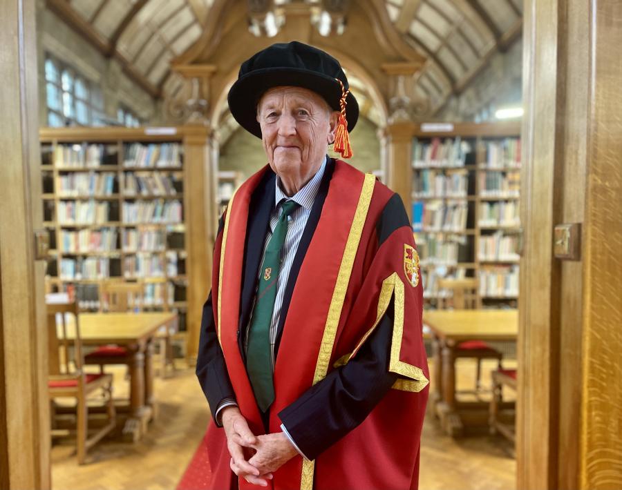 Person yn gwisgo gŵn seremonïol academaidd gyda trim coch ac aur, yn sefyll mewn llyfrgell hanesyddol gyda byrddau pren a silffoedd llyfrau wedi’u llenwi â llyfrau.