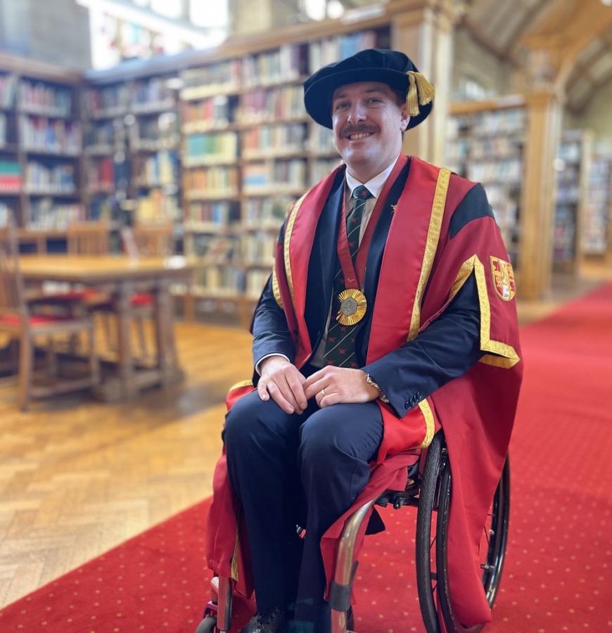 Person in academic ceremonial robes seated in a wheelchair inside a grand library with wooden bookshelves and red carpet.