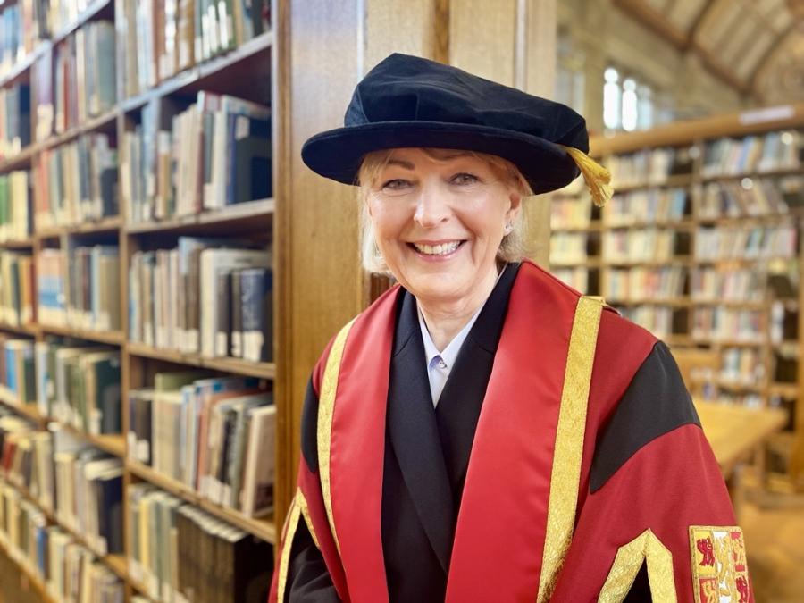 Person yn gwisgo gŵn seremonïol academaidd a chap gyda trim aur, yn sefyll mewn llyfrgell llawn silffoedd llyfrau.