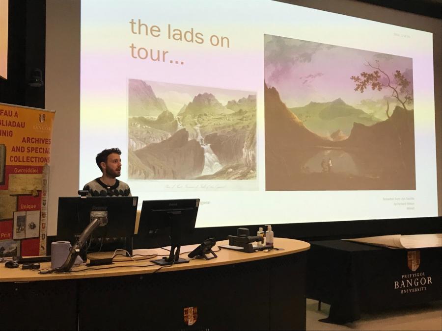 Alex stands behind a lectern presenting. The presentation on screen reads 'the lads on tour...'