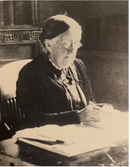Sepia photograph of an elderly woman sat behind a table writing in a large book