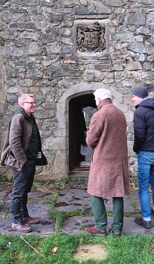 Mae tri dyn yn sefyll mewn sgwrs ac yn gwenu o flaen wal gerrig. Yn y wal, mae drws gyda tharian arfbais wedi'i cherfio uwchben.