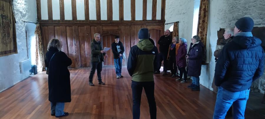 Mae grŵp o bobl yn sefyll mewn neuadd fawr, gyda sgrin bren y tu ôl iddyn nhw. Maen nhw'n gwrando ar ddyn, sy'n darllen o ddarn o bapur.