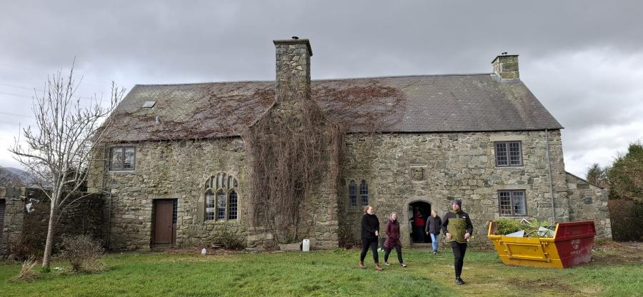 Ffotograff o dŷ carreg hir gyda simnai fawr yng nghanol y ffasâd. Yn y blaendir, mae sgip melyn a sawl person yn cerdded i ffwrdd o'r tŷ.