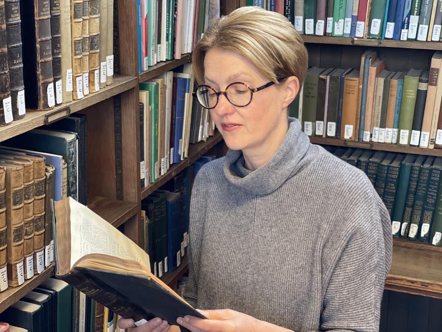 Woman in grey jumper and glasses standing in a library, reading a book. 