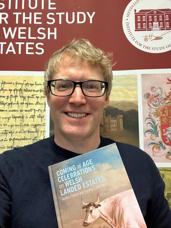 Mae Shaun yn sefyll, yn gwenu, o flaen baner ISWE, yn dal ei lyfr.