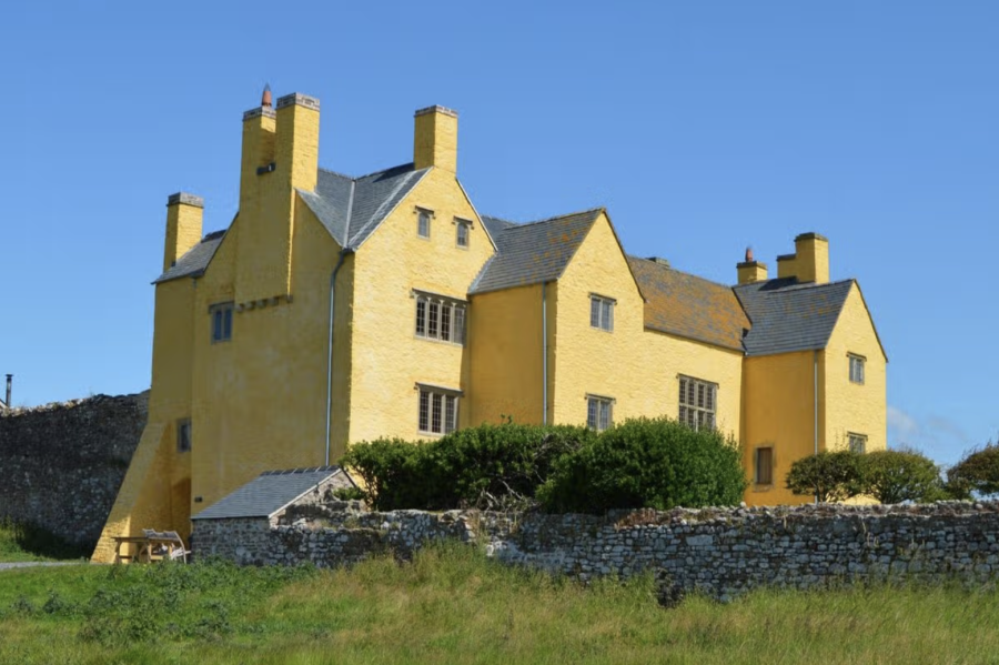 Photograph of a Tudor house with lots of gables and chimneys, with the walls painted bright yellow, set against a blue sky.