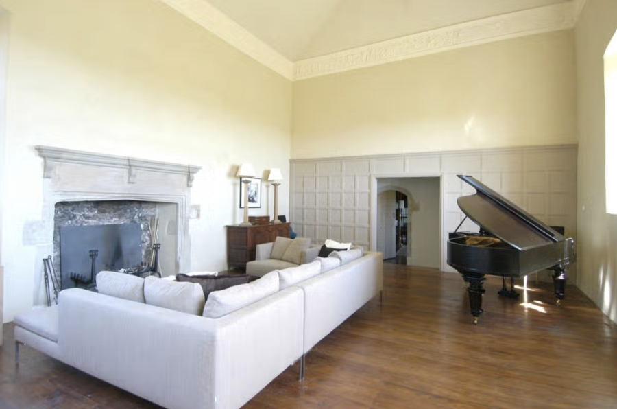 Photograph of a large, tall room with a stone fireplace on one wall and wooden panelling on another. There's a large sofa and a grand piano. 