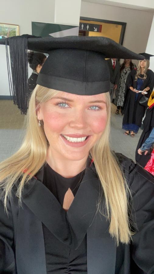Menyw â gwallt melyn mewn gwisg graddio ddu, gyda chap fflat